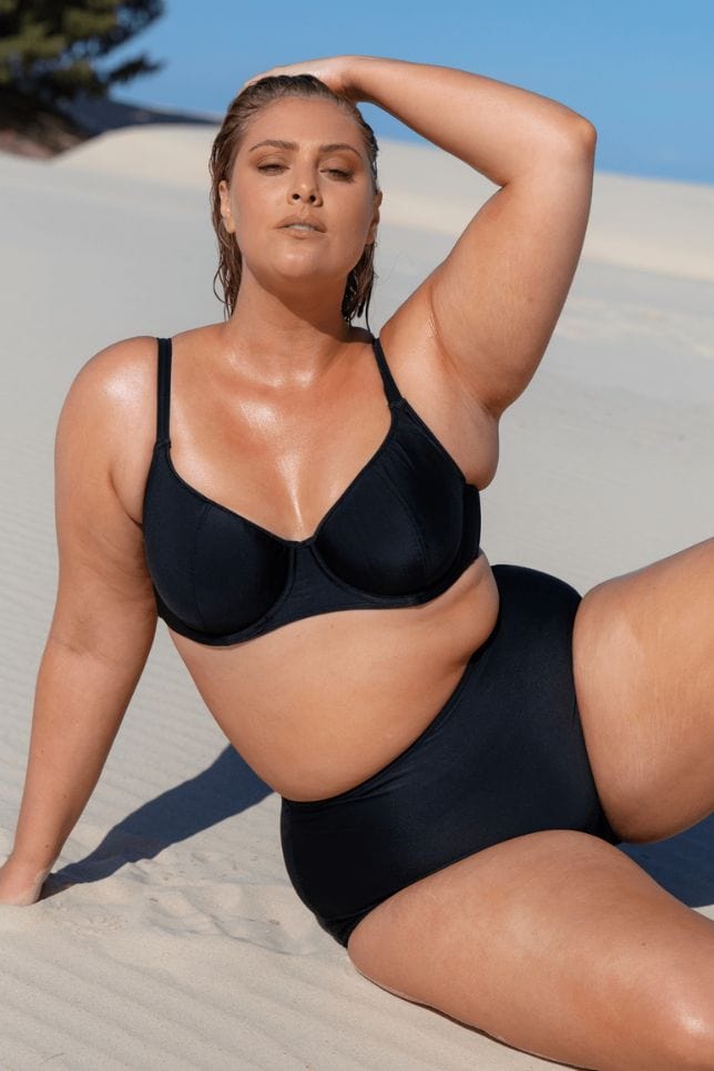 Model sits on the sand wearing a plain black underwire bikini top