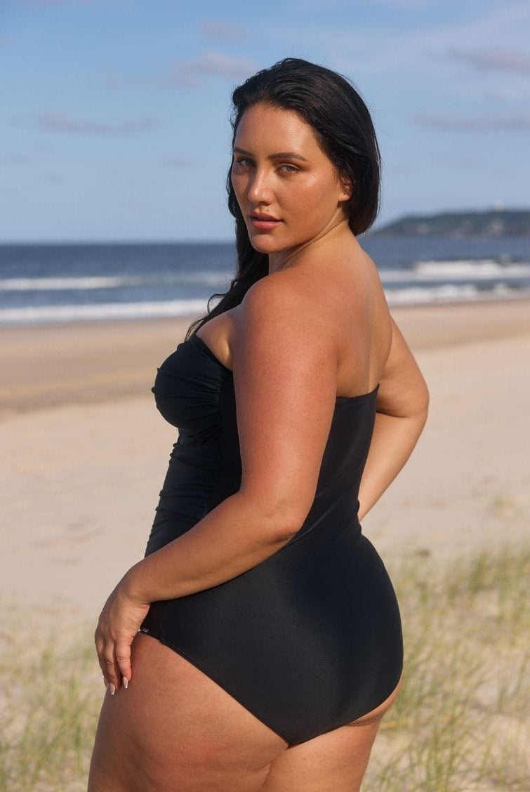A woman wearing a black one-piece swimsuit stands on a sandy beach with the ocean and sky in the background, looking over her shoulder.