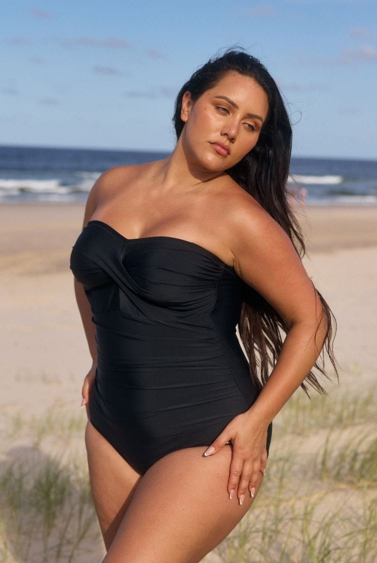 A woman modeling a black strapless one-piece swimsuit on a sandy beach with ocean waves and blue sky in the background.