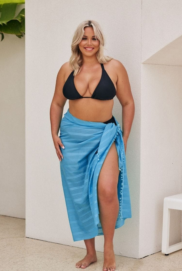 Woman wearing a black bikini top and blue sarong standing indoors.