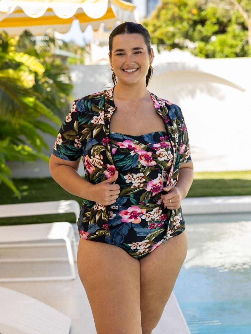 A woman wearing a tropical floral print two-piece swimsuit stands by a swimming pool. The set features a short-sleeve button-up shirt and matching high-waisted briefs with a dark navy background and pink, green, and white floral motifs.