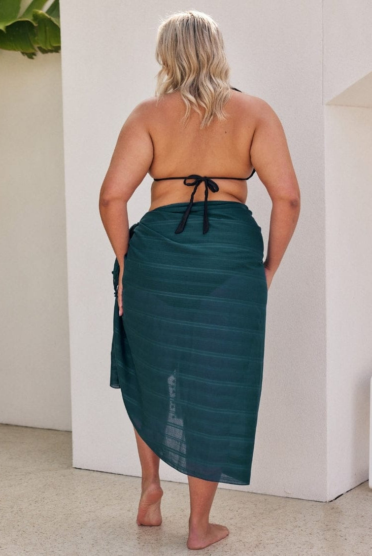 Woman wearing a green sarong standing against a white wall.