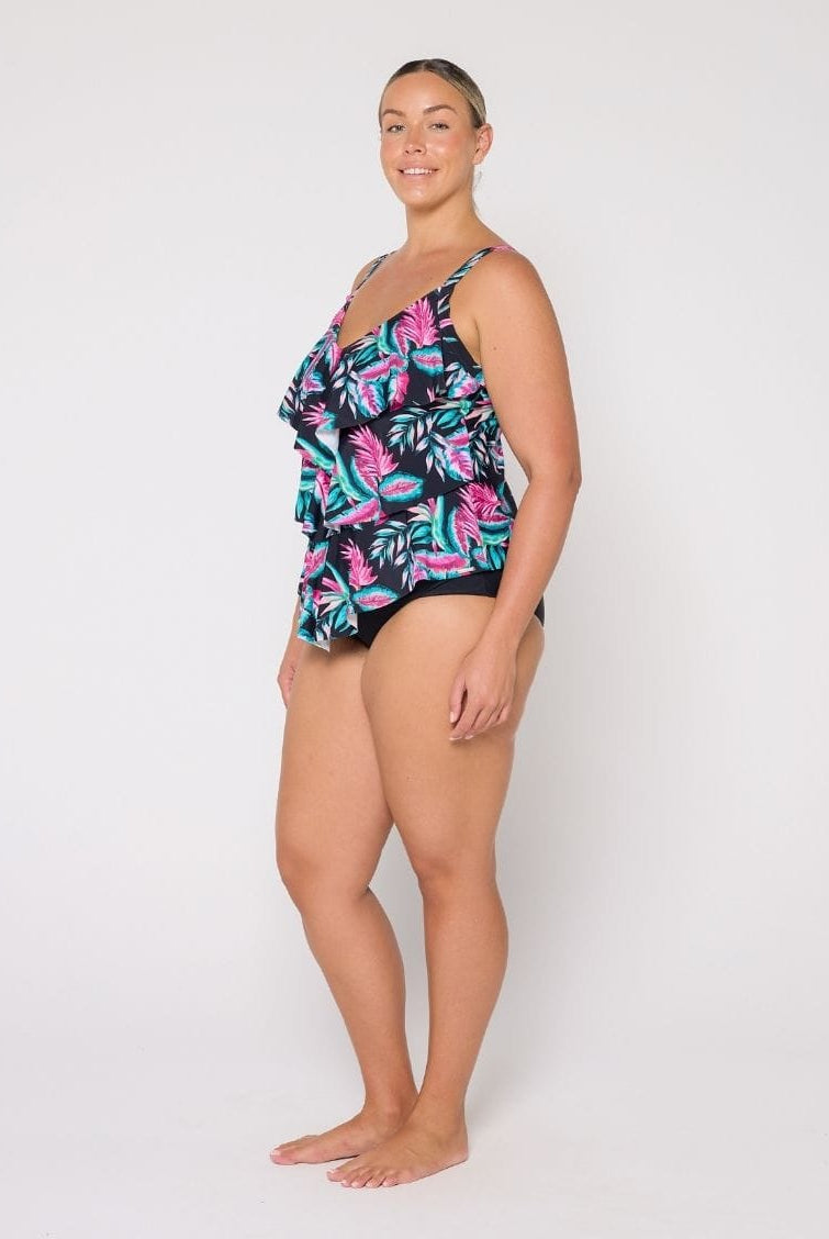 A woman modeling a 3 tier tankini with a tropical leaf pattern in pink, teal, and green on a black background, photographed against a plain white studio backdrop.
