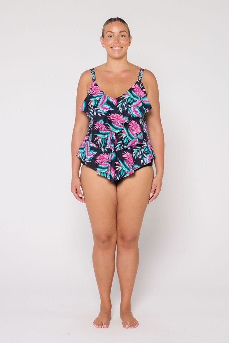 A woman modeling a 3 tier tankini with a tropical palm leaf pattern in pink, teal, and black against a plain white studio background.