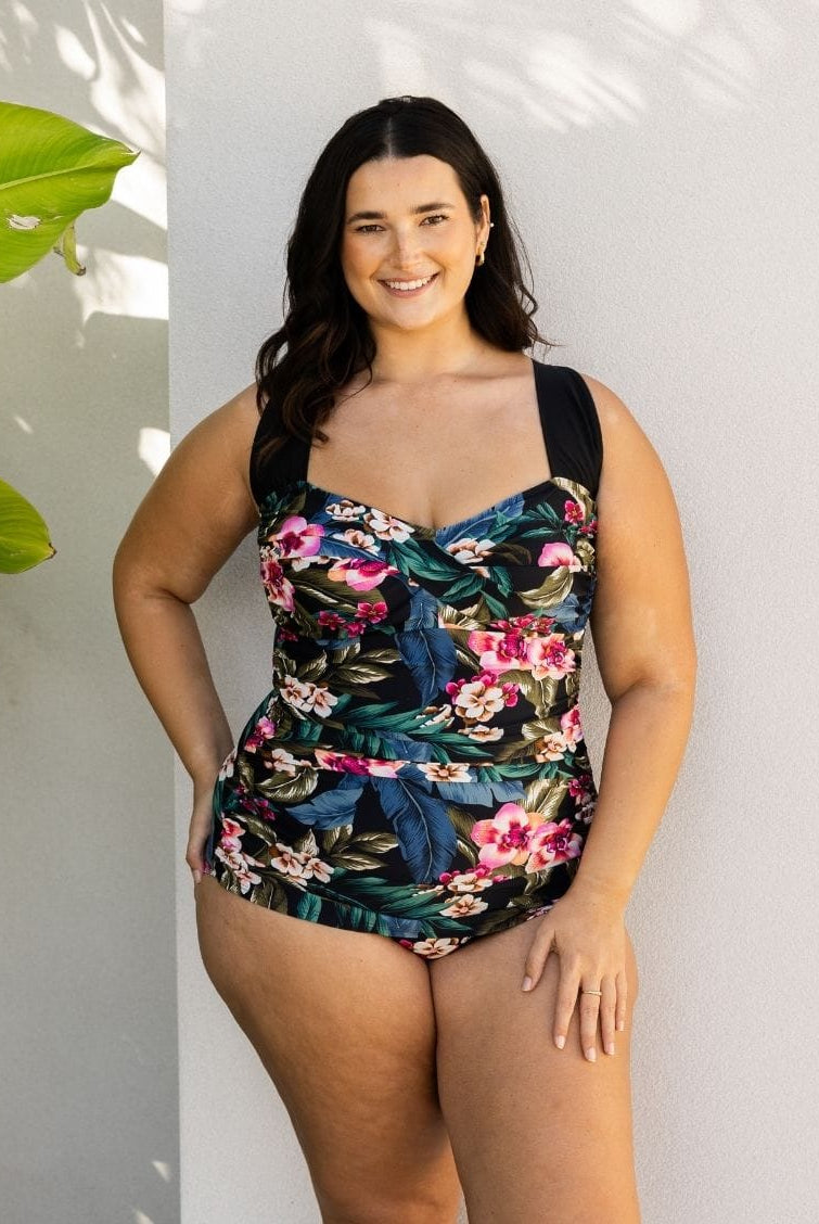 A woman modeling a one-piece swimsuit with a dark tropical floral print, standing against a light gray wall with leaf shadows.