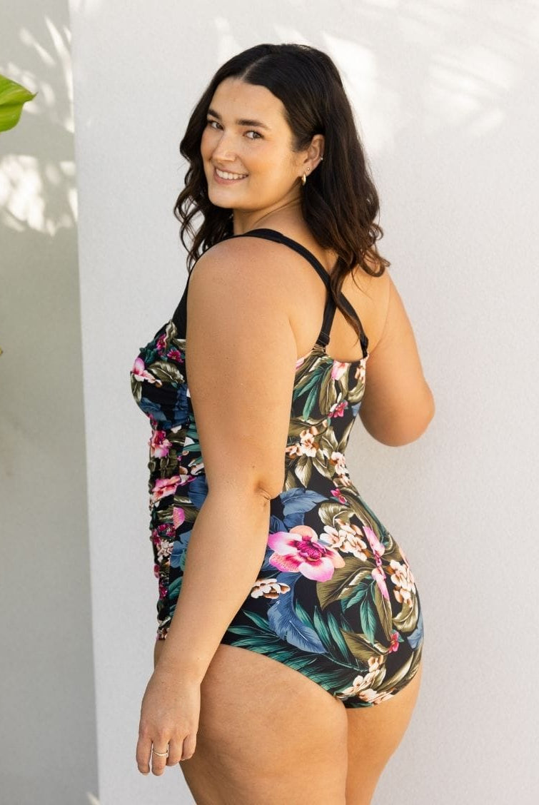 A woman modeling a one-piece swimsuit with a tropical floral print, photographed against a white wall with soft leaf shadows.