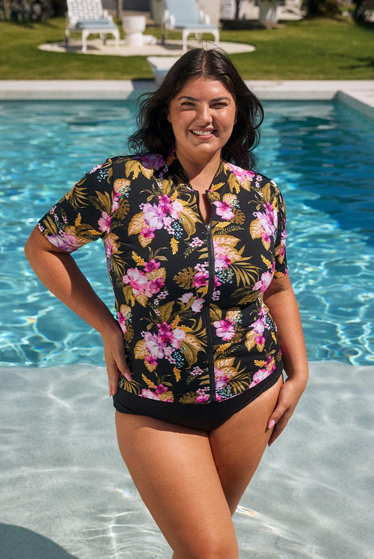 Brunette model wears stunning pink and gold floral short sleeve rash vest in chlorine resistant fabric with front zip