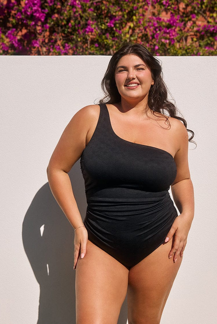 Woman wearing a black one-shoulder swimsuit standing against a light background with purple flowers.
