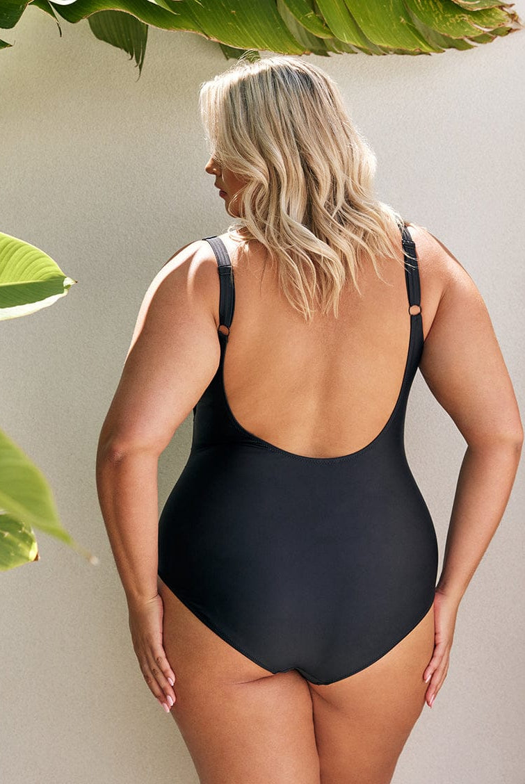 back of model standing in front of a white wall wearing a black one piece swimsuit