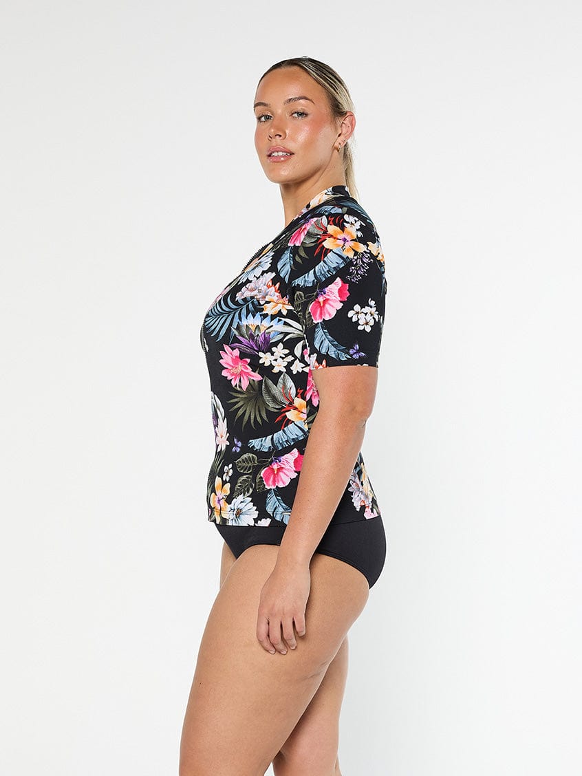Blonde model shows the side of pool proof rash vest with short sleeve in a black based floral print