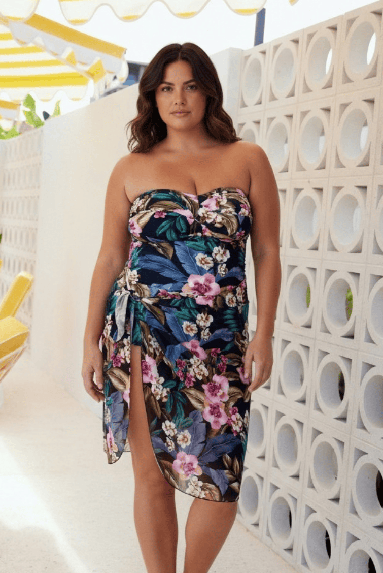 A woman models a strapless floral wrap dress with a sweetheart neckline and a thigh-high slit. The dress features a vibrant tropical floral pattern in shades of navy, teal, pink, and green.