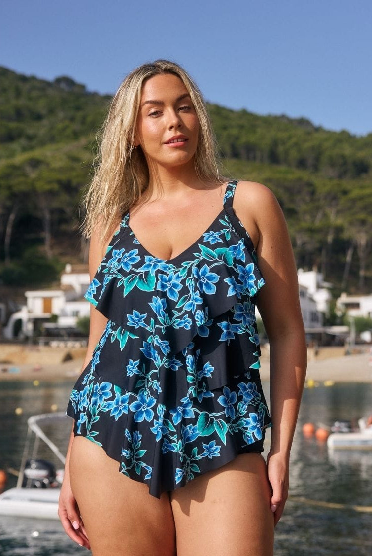 A woman wearing a black one-piece swimsuit with a blue and green floral print stands outdoors in front of a beach and water backdrop.