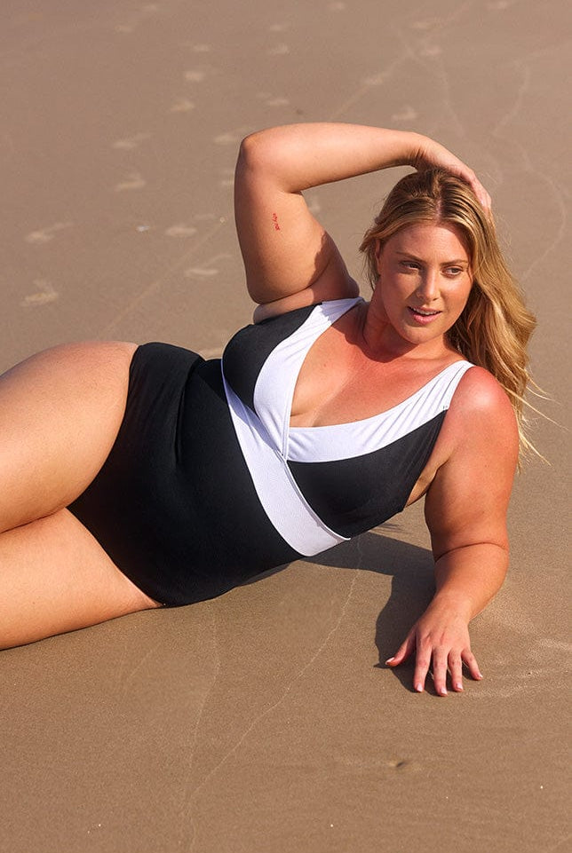 Blonde model lays on the sand, wearing a black v-neck one piece swimsuit with a white trim.
