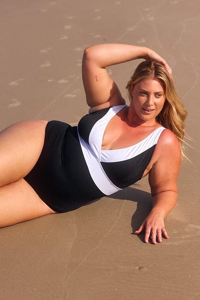 Blonde model lays on the sand, wearing a black v-neck one piece swimsuit with a white trim.