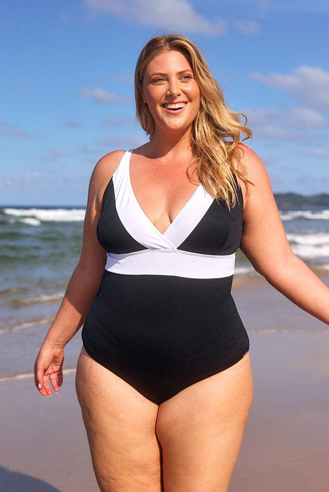 Blonde model at the beach, smiling at the camera, wearing a black plunge one piece swimsuit with a white trim.