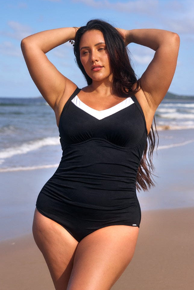 Brunette model stands at the beach, wearing a black underwire v-neck one piece with a white trim. The supportive cup sized swimsuit has ruching down the torso for a slimming effect.