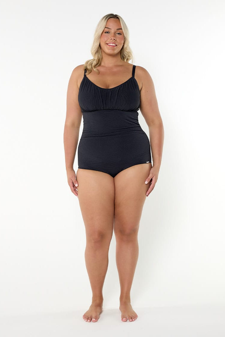 A woman modeling a black one-piece swimsuit with thin shoulder straps and a gathered sweetheart neckline, standing against a plain white studio background.