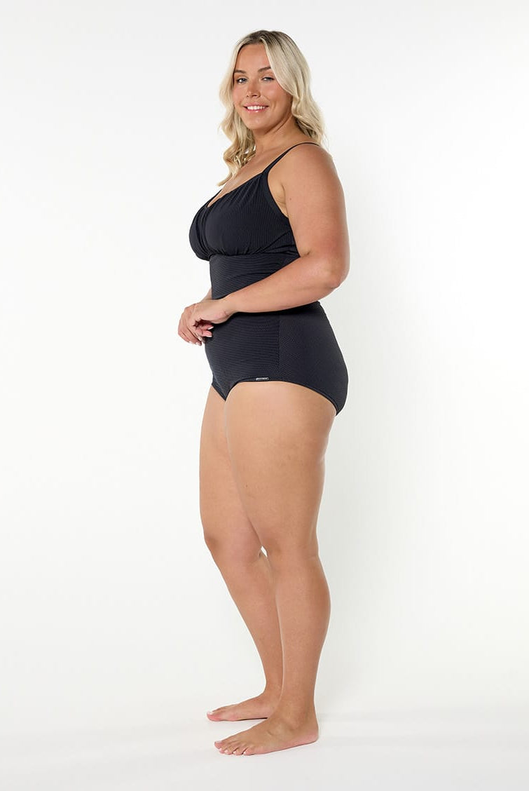 A woman modeling a black one-piece swimsuit against a plain white studio background, shown in a side profile view.