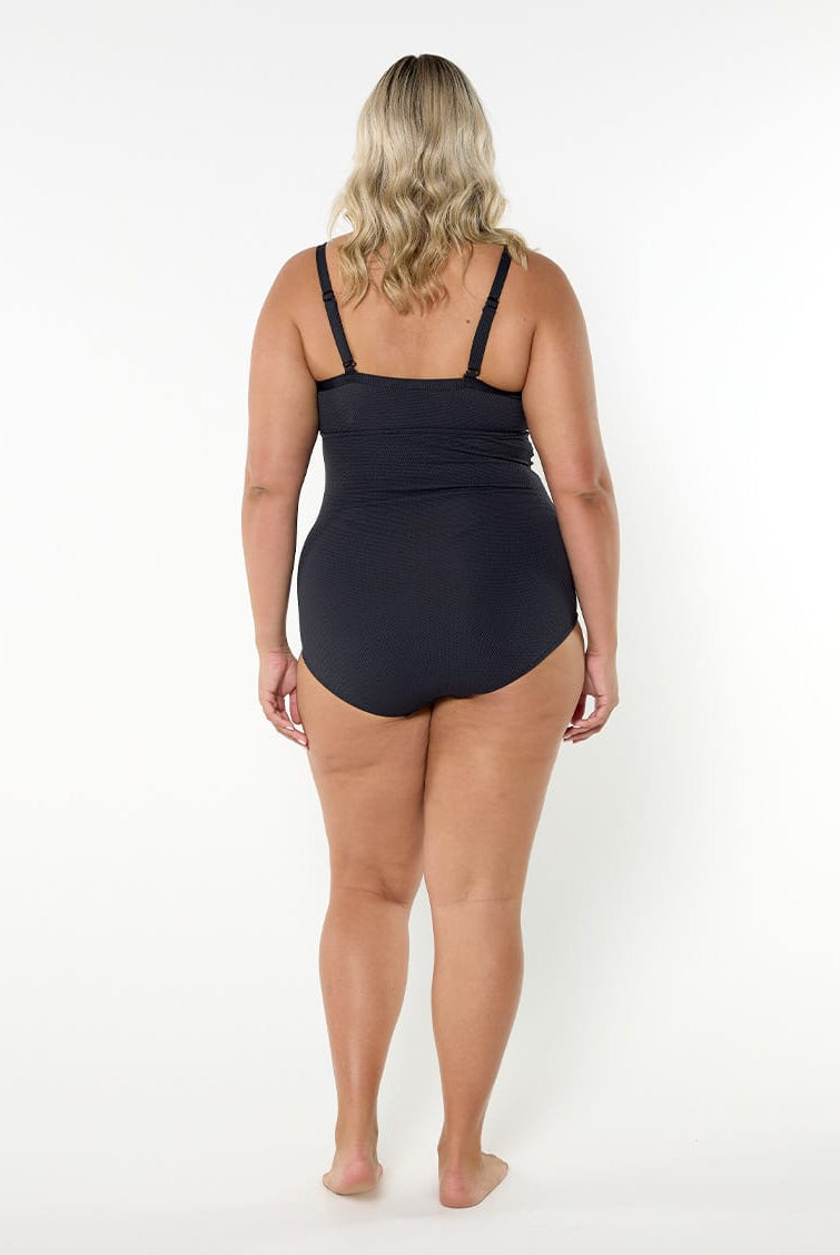 A woman viewed from the back wearing a black one-piece swimsuit, standing against a plain white studio background.