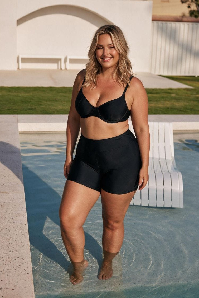 A woman modeling a black two-piece swimwear set consisting of a bandeau-style bikini top and high-waisted shorts, standing in shallow water next to a pool in an outdoor backyard setting.