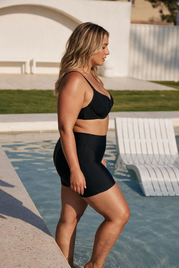 A woman wearing a black two-piece swimwear set consisting of a sports bra and high-waisted shapewear briefs, standing by a swimming pool in a sunny outdoor setting.