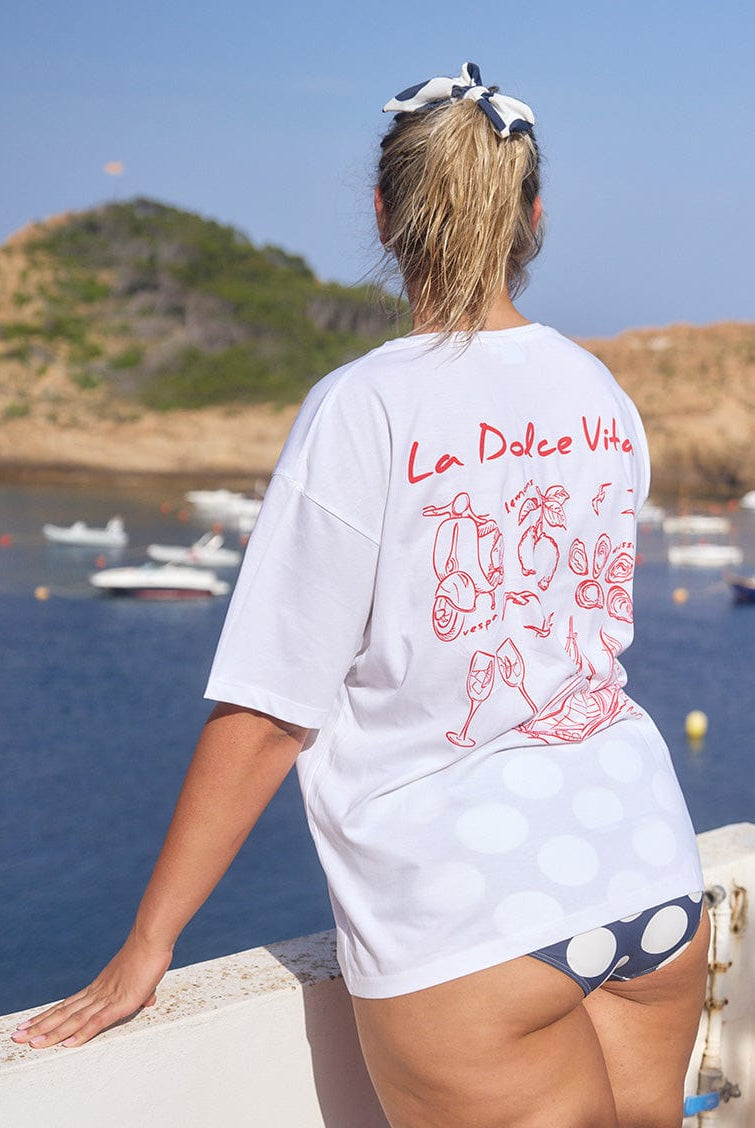 Woman wearing a white t-shirt with 'La Dolce Vita' design by a waterfront with boats and green hills.