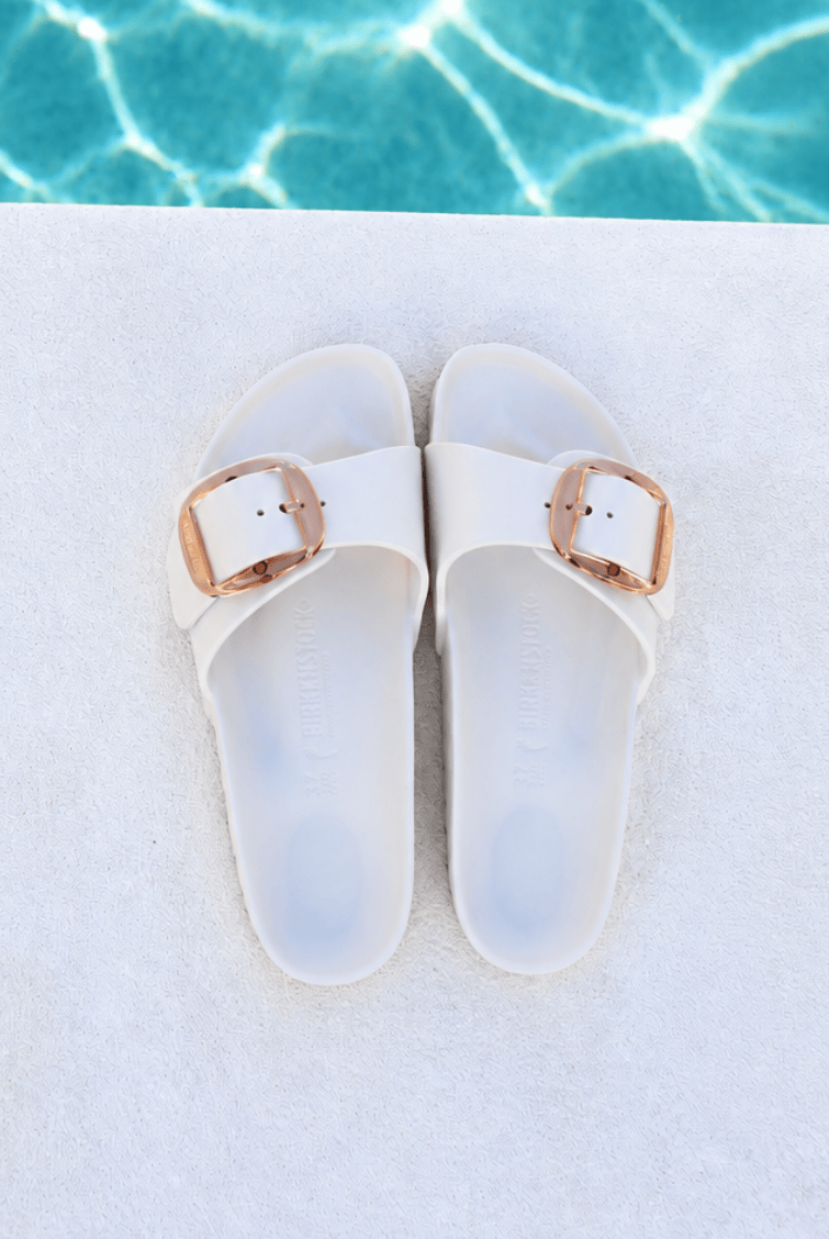 A pair of white slide sandals with metallic rose gold buckle straps, laid flat on a white textured surface with a swimming pool background visible at the top.