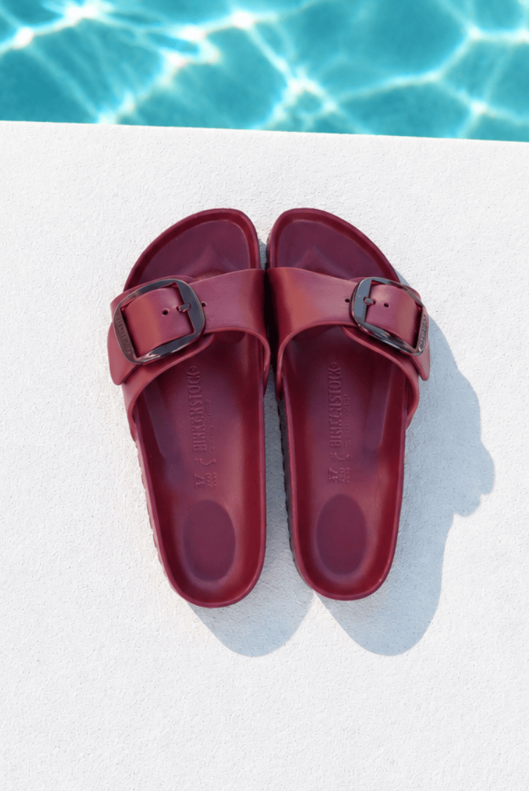 A pair of burgundy double-strap slide sandals with dark metal buckles, laid flat on a white deck with a swimming pool background visible at the top.