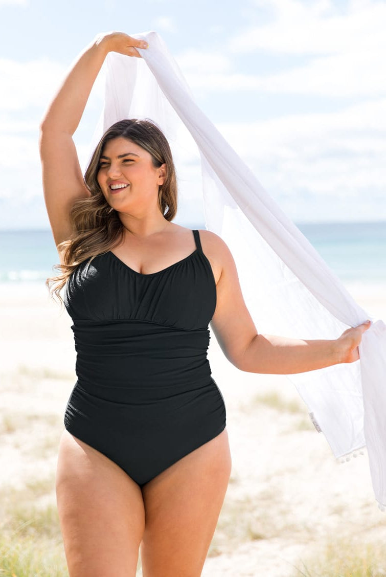 A woman stands on a sandy beach wearing a black underwire one-piece swimsuit and holding a white scarf overhead.