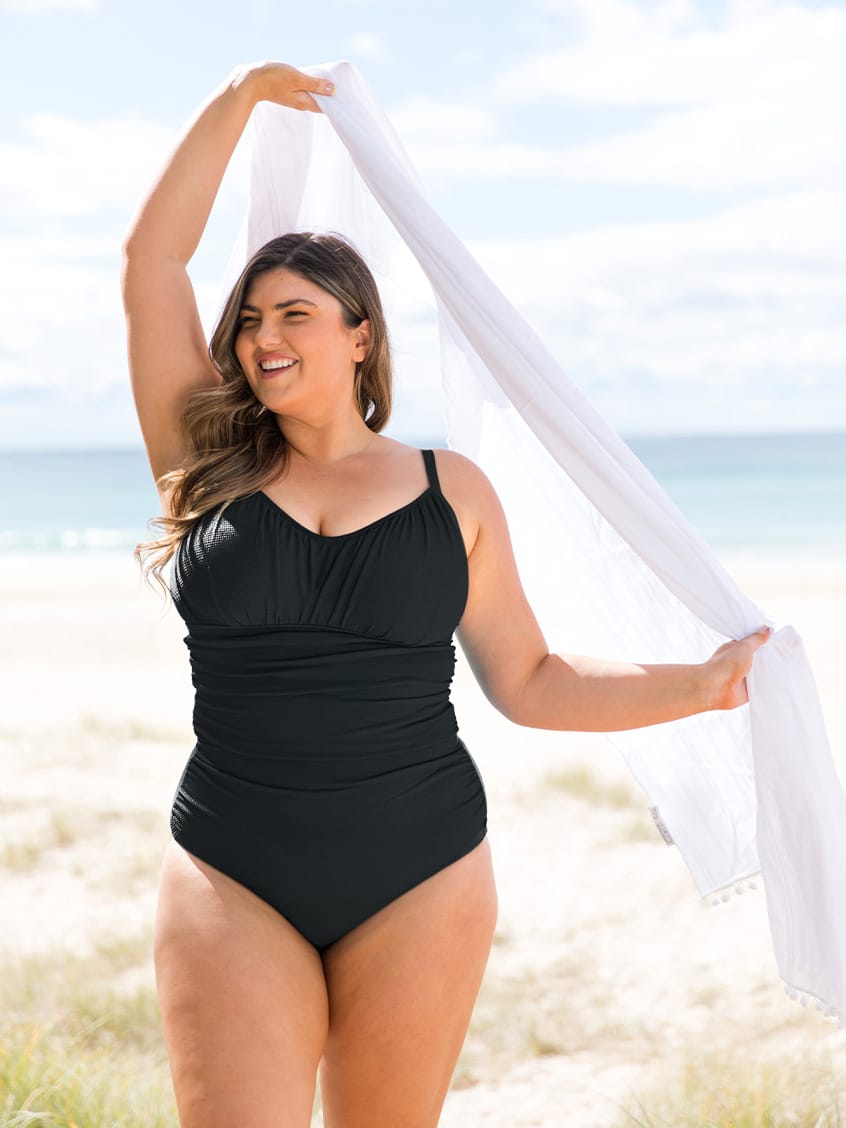 A woman stands on a sandy beach wearing a black underwire one-piece swimsuit and holding a white scarf overhead.