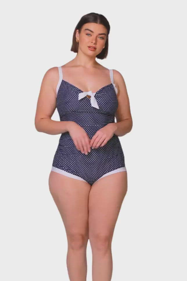 brunette model wearing navy and white dots boyleg one piece swimsuit
