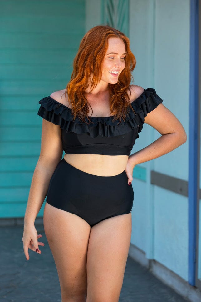 Model wearing a plain black bikini top. The top has two frills along the bustline and is worn off the shoulder