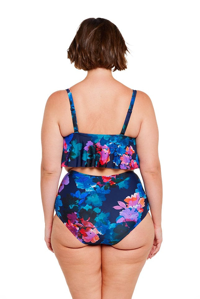 Brunette model shows back of navy floral frill bikini top and high waisted pant