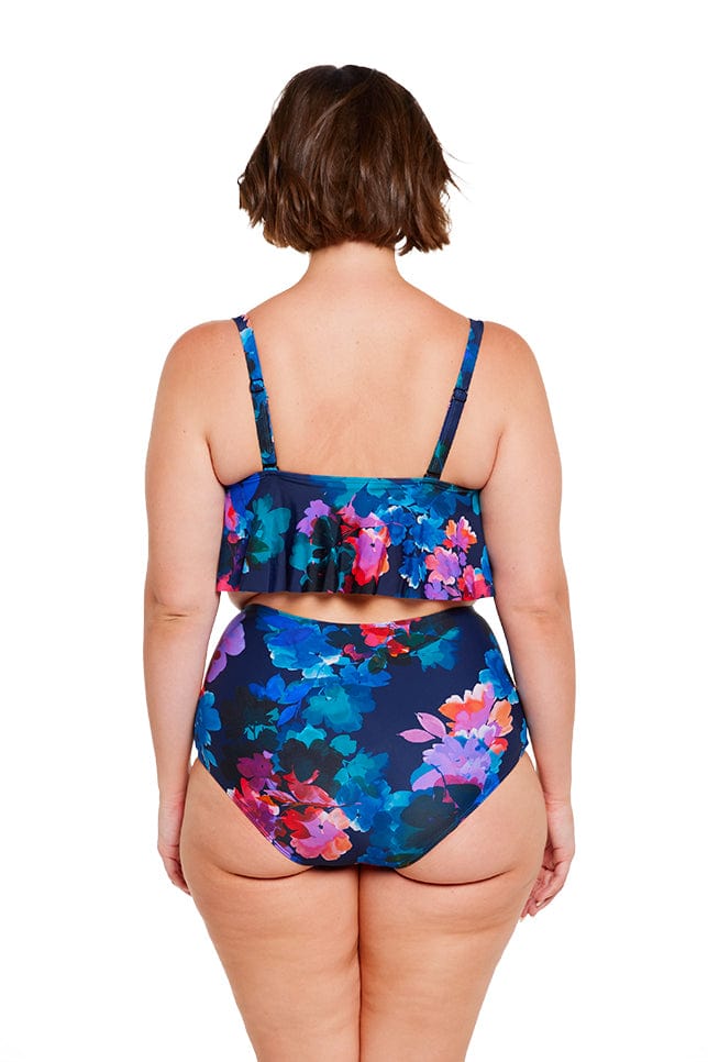 Brunette model shows back of navy floral frill bikini top and high waisted pant