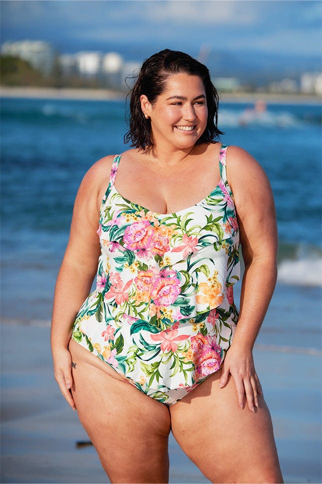 brunette plus size model wears white floral one piece with 3 tier ruffle details at the beach