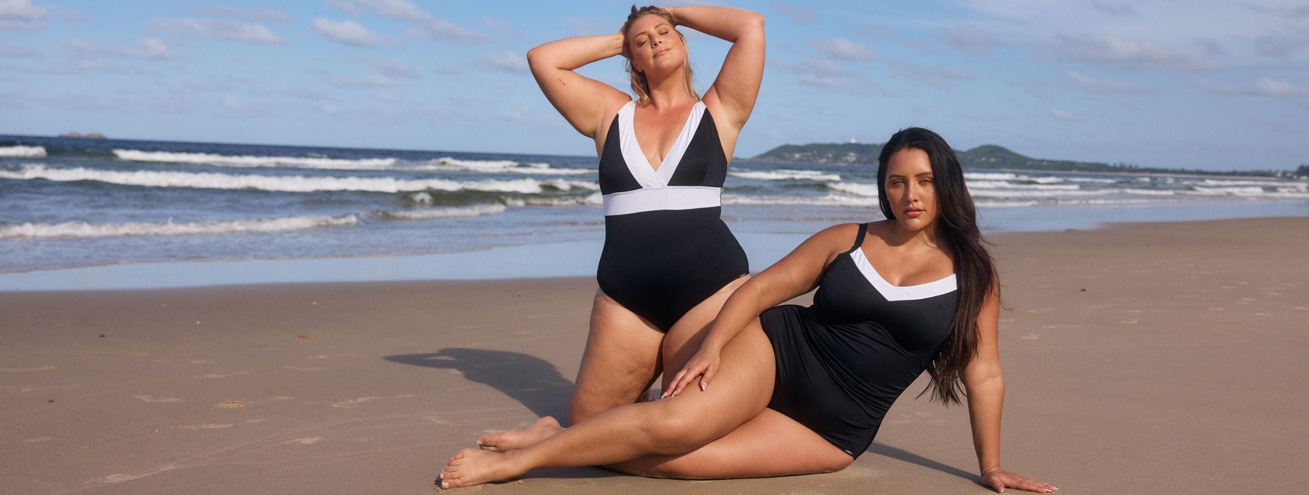 Two women pose on the beach wearing black and white one piece swimsuits 