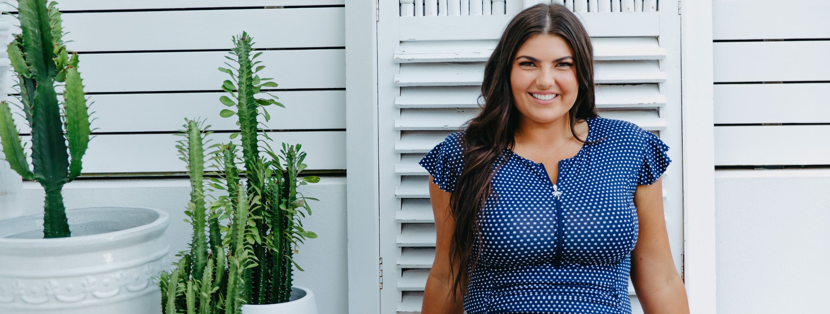 Woman with long dark brown hair wears navy and white polkadot swimsuit with zipup and frill sleeves