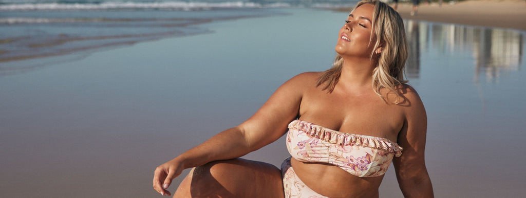 Blonde woman sits on the beach wearing pale pink floral strapless bikini top and high waisted pants