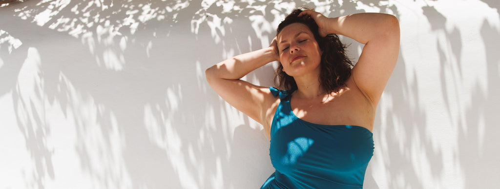 Woman with dark brown curly hair wears a Deep Teal metallic one shoulder one piece