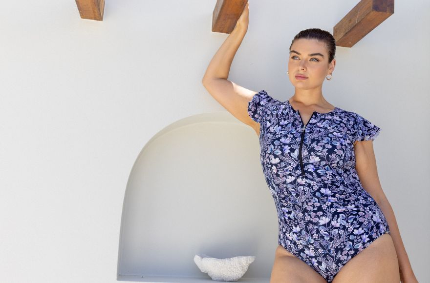 Brunette woman poses by a white wall wearing navy floral frill sleeve zip up one piece swimsuit