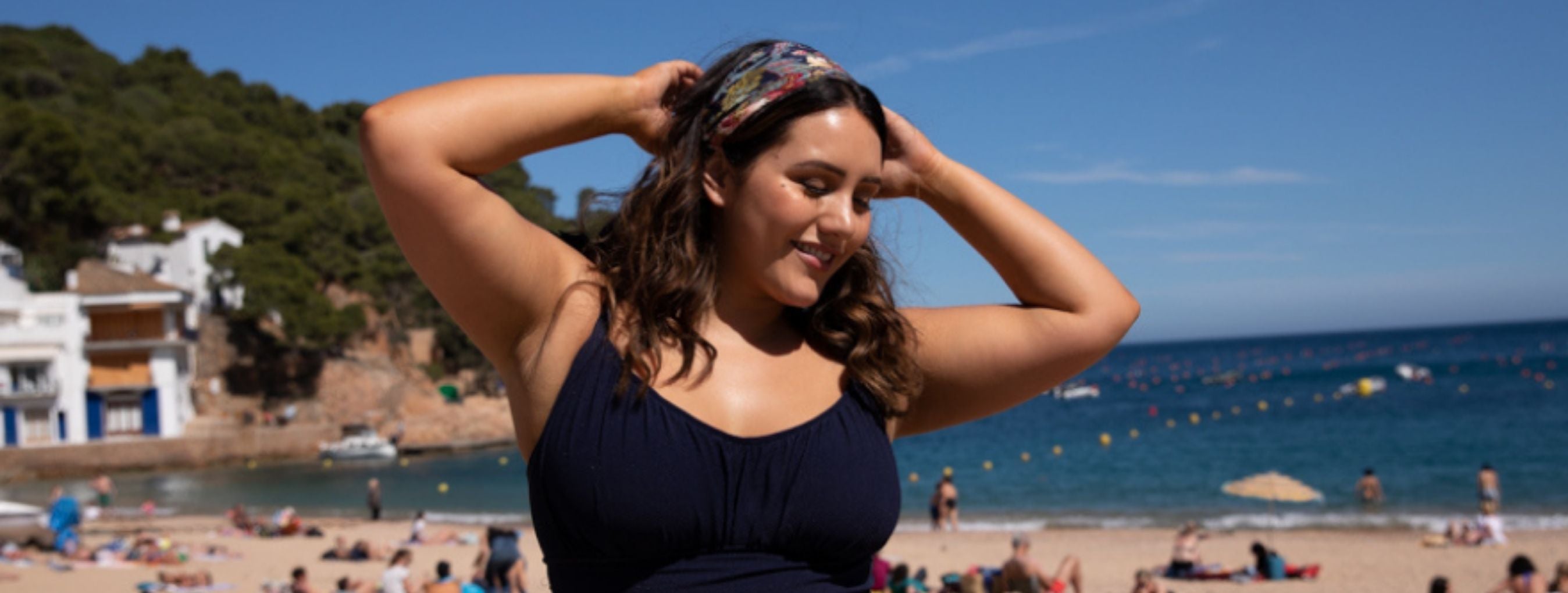 Brunette woman poses on the beach wearing Honey Comb black one piece. She has bronzed glowing skin from applying self tan.
