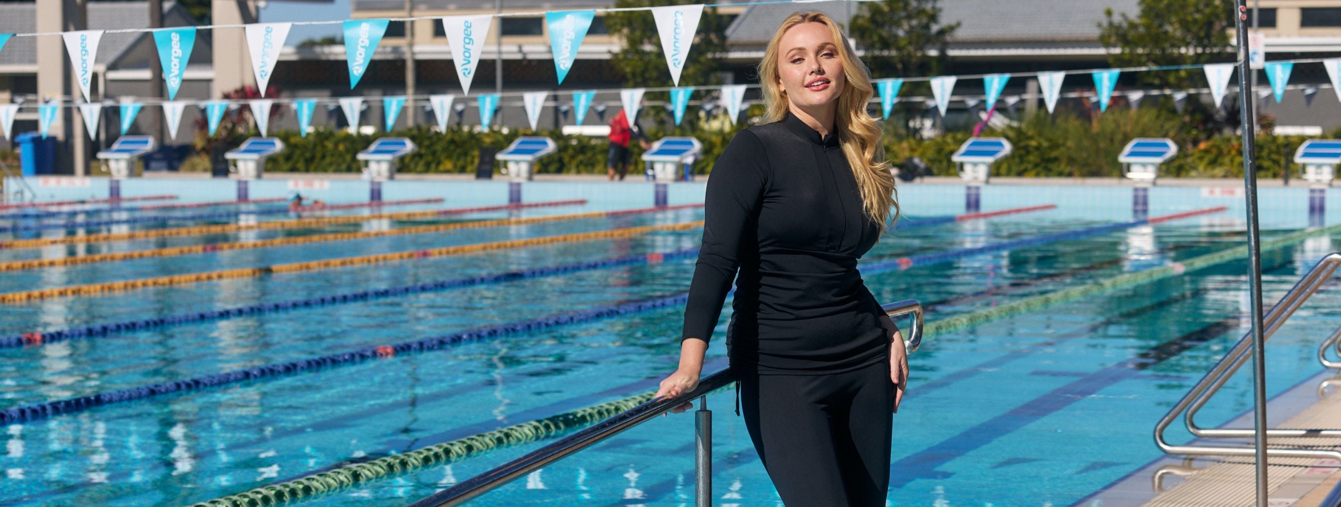 Woman with long blonde hair poses by the pool wearing black long sleeve rash vest with full length swim leggings. 