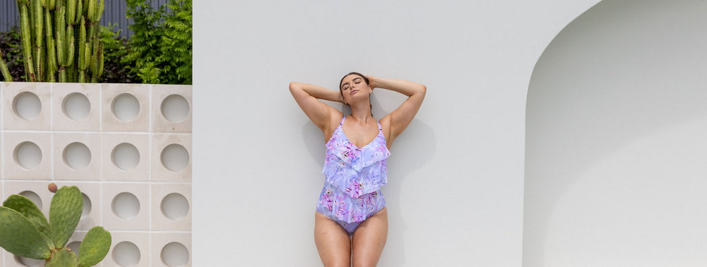 Brunette model poses by white wall wearing Lilac Florence 3 tier one piece swimsuit