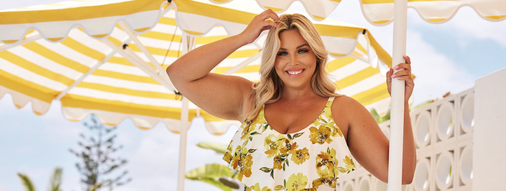 Woman with blonde hair stands under a yellow and white striped umbrella wearing a white one piece swimsuit with yellow flowers