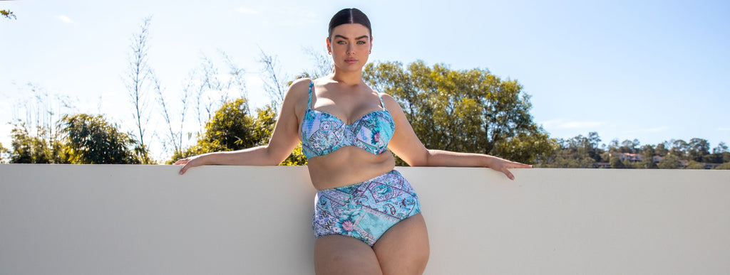 Woman with slicked back brown hair poses against a white wall wearing Whitehaven blue patchwork underwire bikini top and high waisted pant 
