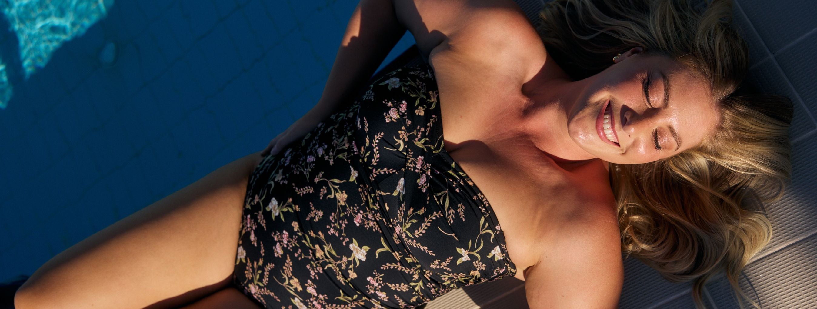 Woman with blonde hair reclines by the pool wearing a black floral strapless swimsuit