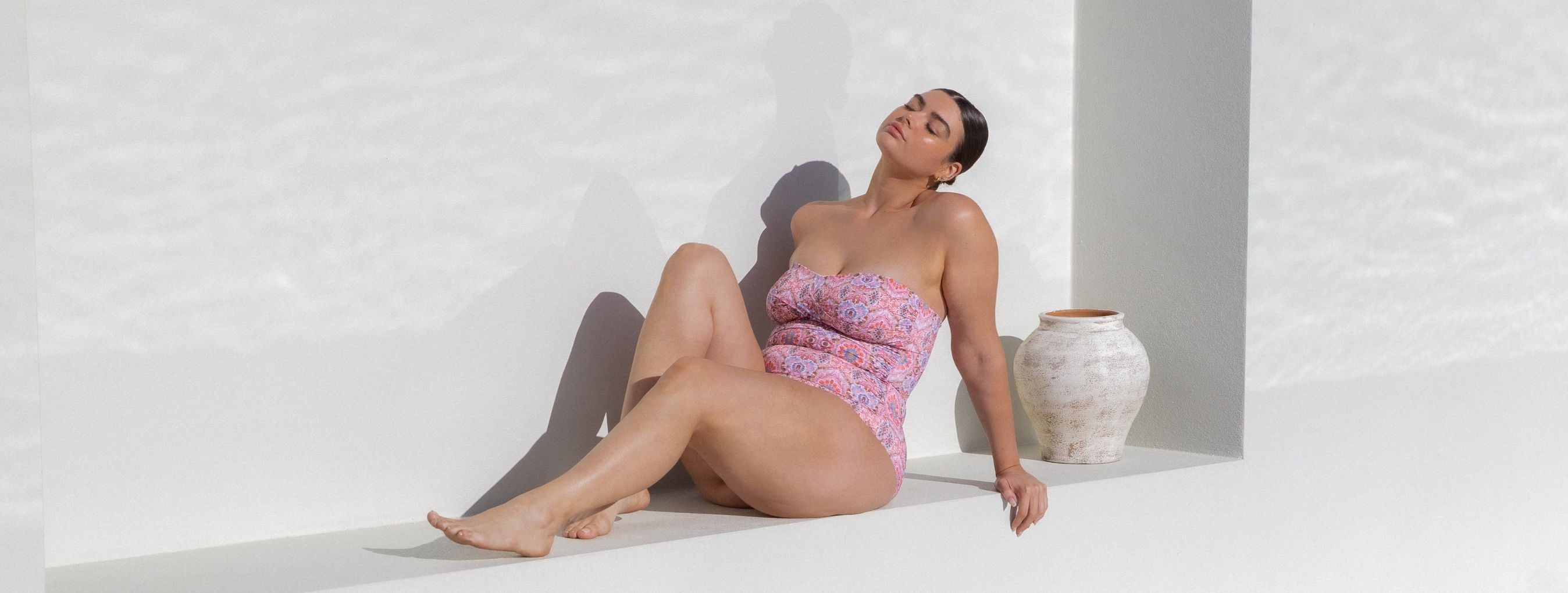 Brunette woman poses on a white bench wearing Amalfi Pink boho print strapless twist front bandeau one piece swimsuit
