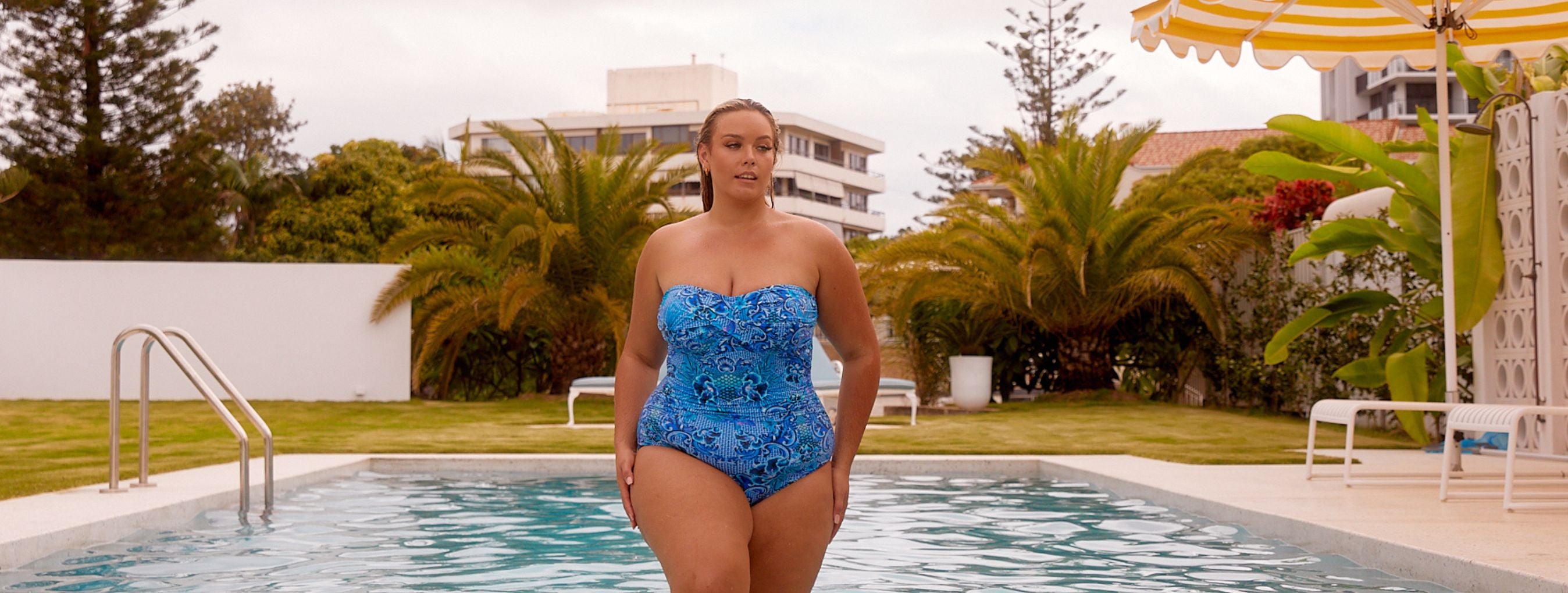 Woman with slicked back blonde hair steps out of the pool wearing a blue printed strapless swimsuit