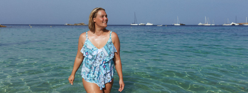 Woman with short blonde hair wades through the water wearing Whitehaven blue patchwork print 3 tier tankini top and matching pants. 