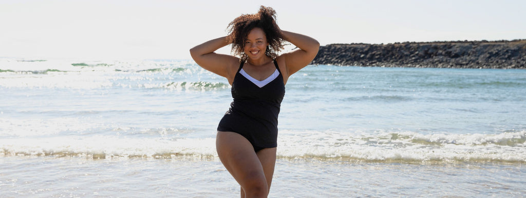 Woman with curly brown hair poses on the beach wearing black tankini top with white v neck trim